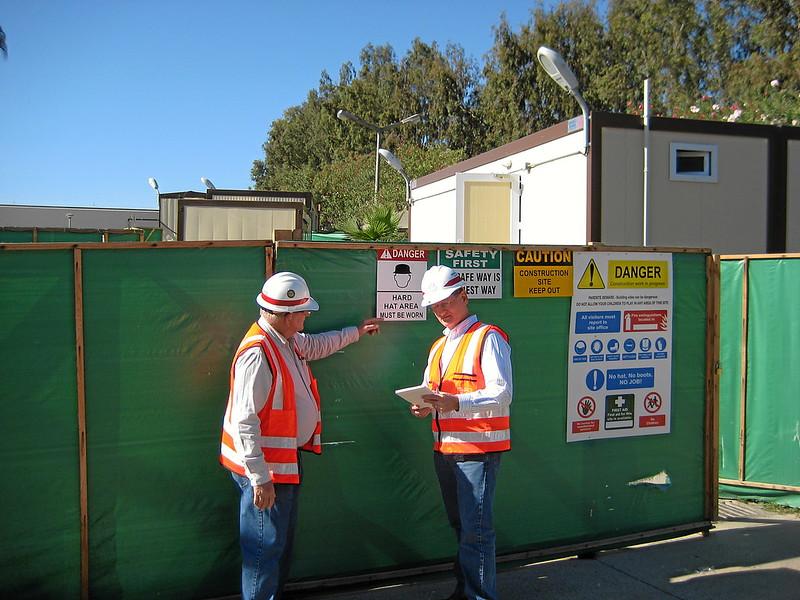 Two men in hard hats and high-visibility safety vests point to safety signs on a temporary construction fence.