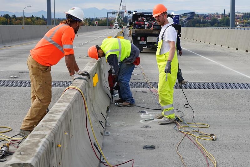 Three workers in high-visibility safety gear lean over concrete barriers on a road construction site.
