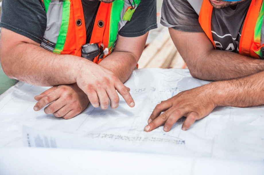 Two people in high visibility safety vests point at building blueprints on a construction site.