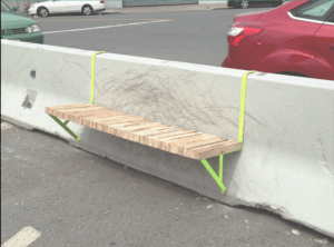 A wooden and metal bench is clipped to the top of a concrete jersey barrier