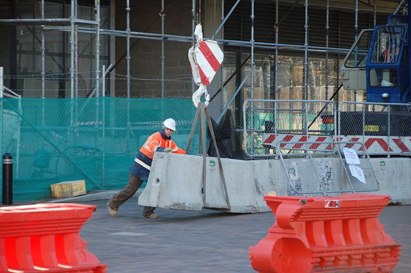 A worker in a hard hat and safety vest pushes a concrete barrier that is held above a the ground by a crane and a heavy-duty straps.