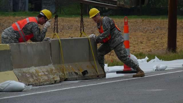Two army members in camouflage, hard hats, and safety vests place a concrete jersey barrier that is suspended by straps from a crane.