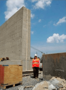 A person wearing a safety vest stands next to a T-wall to illustrate its relative height and thickness.