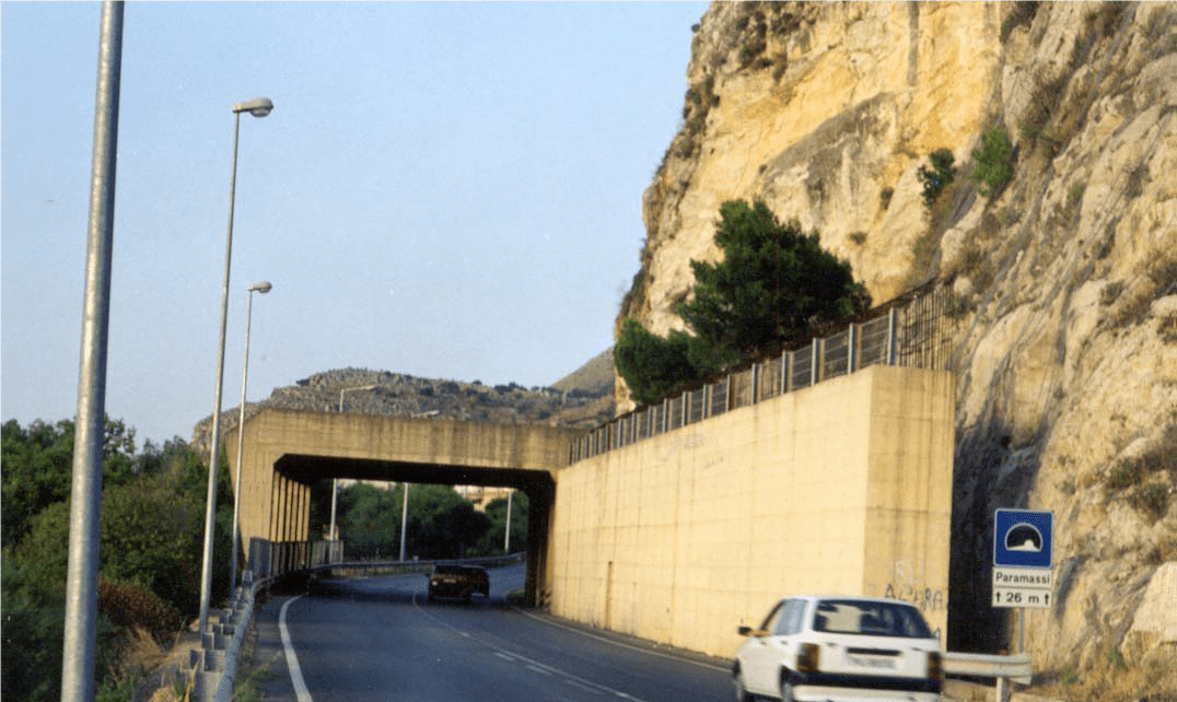 A tall concrete barrier wall stands between a cliff and a roadway to stop rockfall