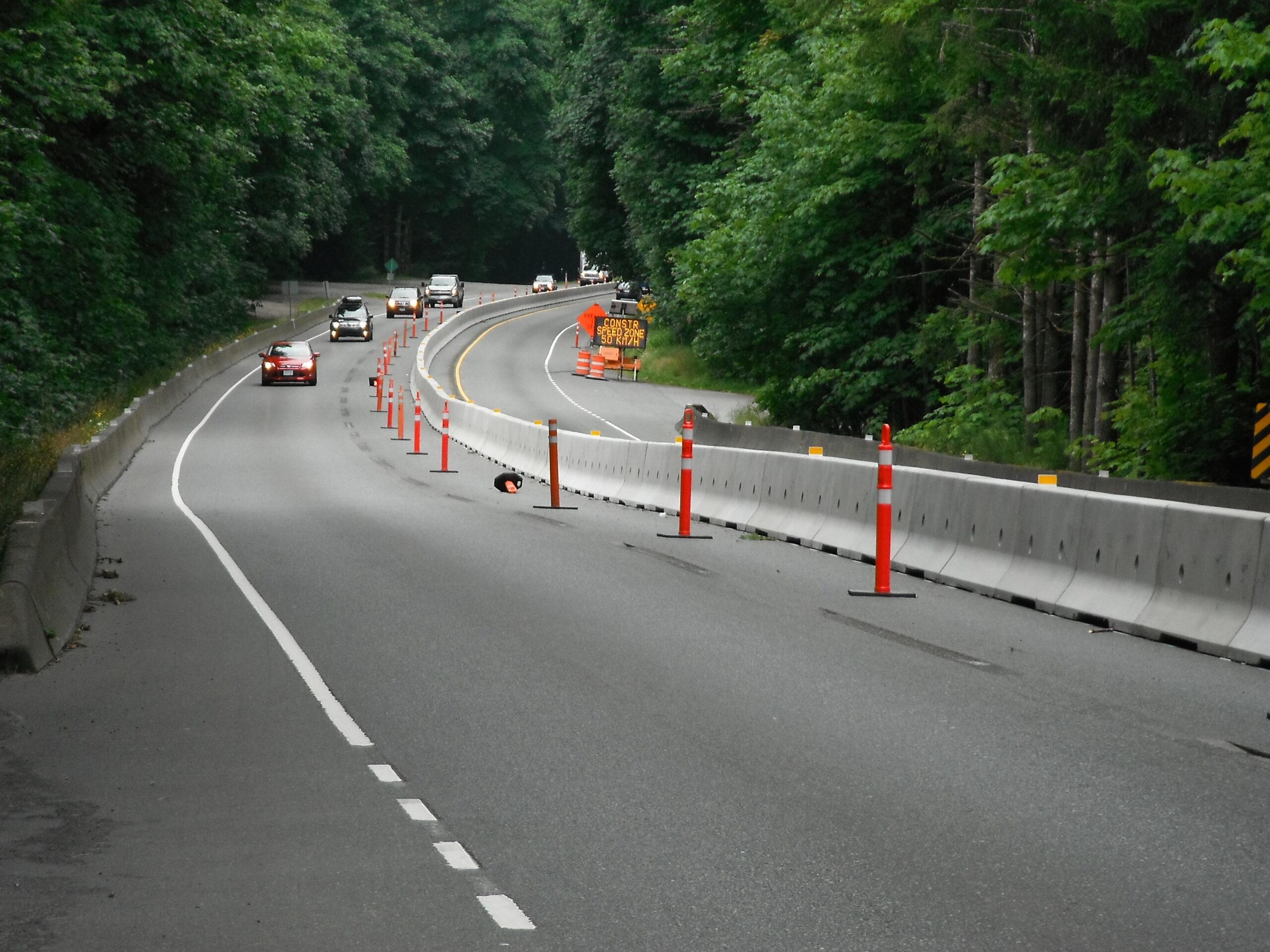 Orange safety cones line a concrete barrier wall along a curved roadway as a line of cars drives past in the adjacent lane.